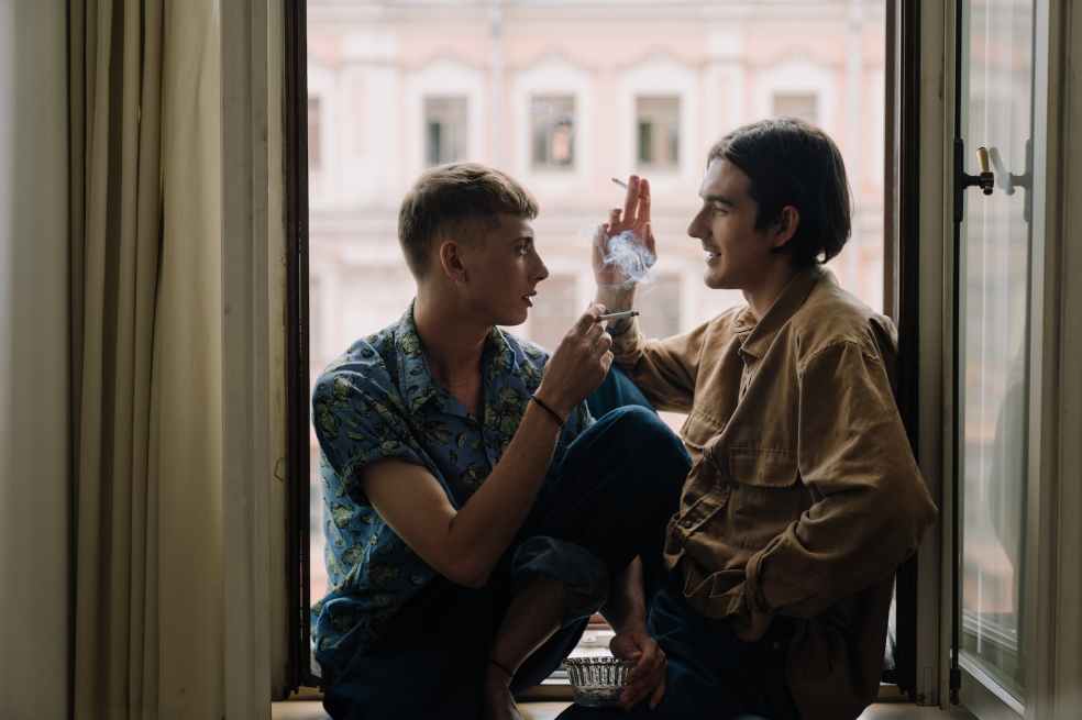 two men facing each other smoking while sitting on the windowsill